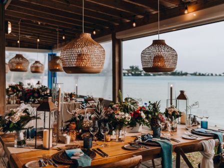 A beautifully decorated dining area with hanging lights and floral centerpieces, set beside a serene waterfront view.