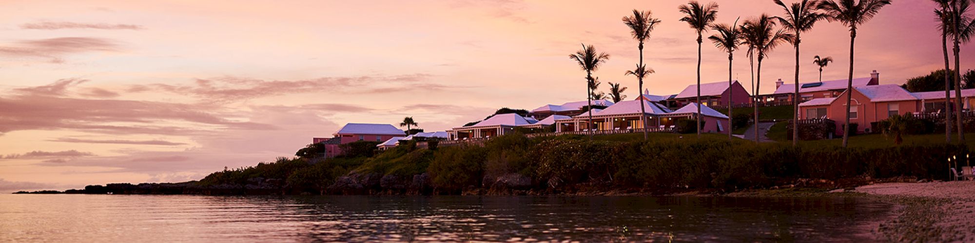 A serene coastal scene at sunset, with colorful skies, palm trees, and cozy houses along the shoreline, reflecting on the calm waters.