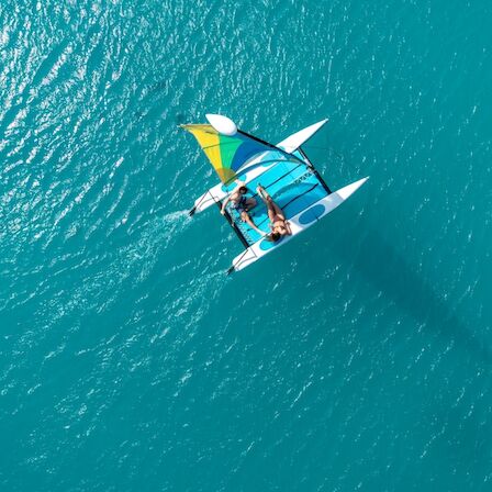 Two people sail a small catamaran with colorful sails on turquoise open water, casting a long shadow beneath them, from a top-down view.