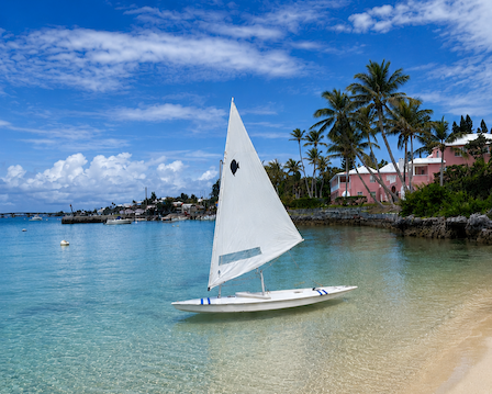 A white sailboat with a blue accent floats in clear tropical water beside palm-lined buildings under a blue sky.