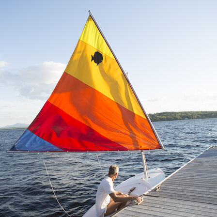 A person sits at the edge of a dock with a small sailboat, its bright red, orange, and yellow sail catching the breeze on a calm lake.