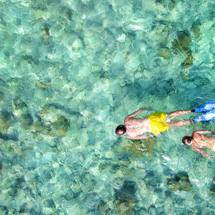Two people snorkeling or swimming in clear turquoise water, wearing bright swim trunks, fins, and floating above a rocky seabed.