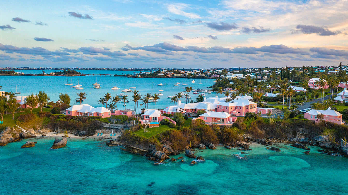 A scenic coastal view featuring turquoise waters, rocky shores, and a collection of pastel-colored houses surrounded by greenery, with boats in the distance.