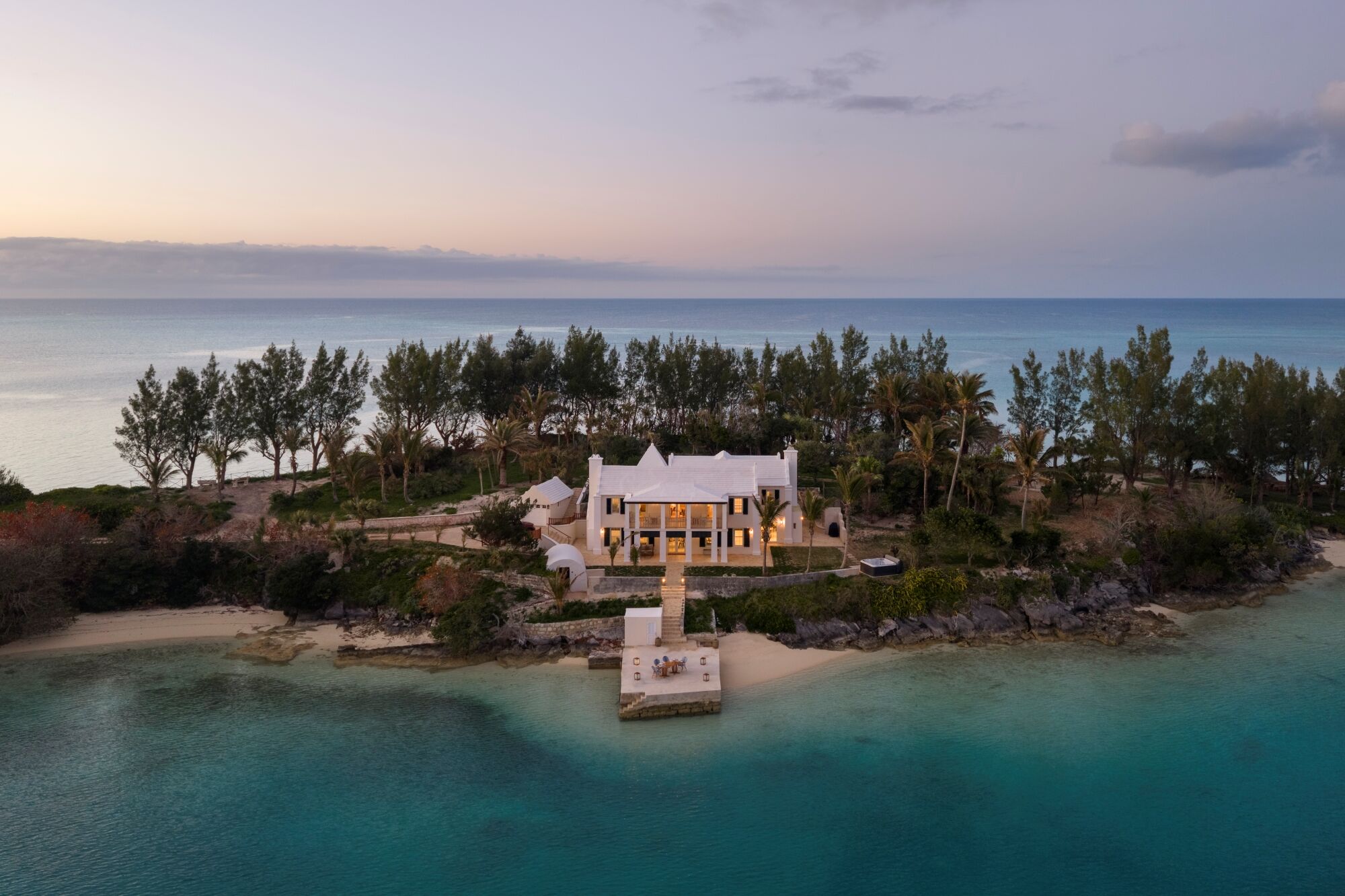 A white coastal villa sits on a rocky shore, surrounded by trees, with turquoise water and a small pier extending into the sea at sunset.