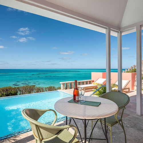 A balcony with a round table and chairs overlooks a turquoise pool and ocean, under a bright blue sky with a rocky coastline in the distance.