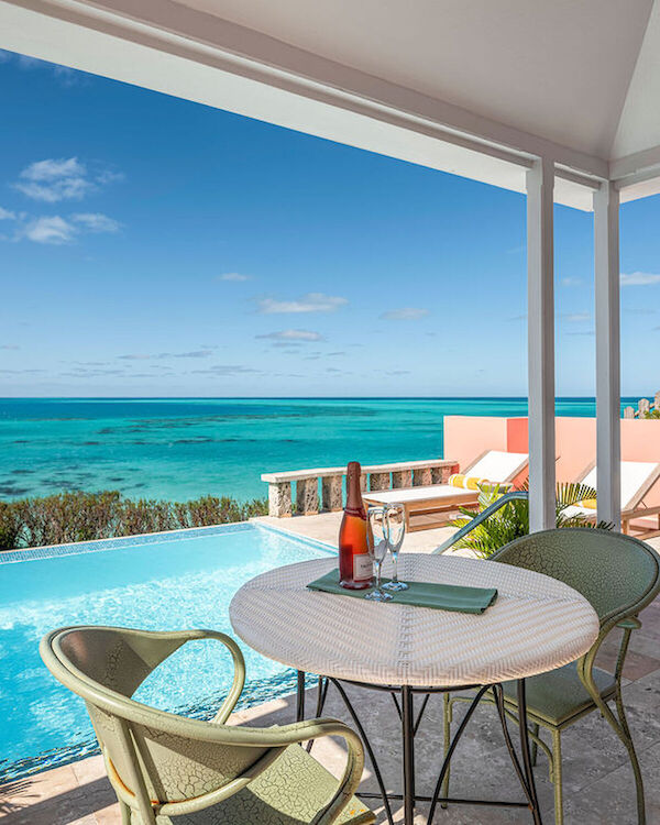 A balcony with a round table and chairs overlooks a turquoise pool and ocean, under a bright blue sky with a rocky coastline in the distance.