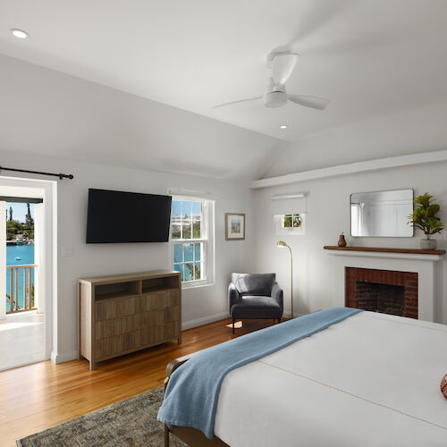 A bright hotel-like bedroom with a bed, fireplace, flat-screen TV, seating area, wooden floor, and a door to a balcony overlooking water.