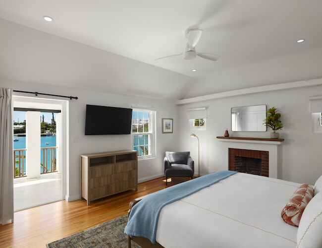 A bright hotel-like bedroom with a bed, fireplace, flat-screen TV, seating area, wooden floor, and a door to a balcony overlooking water.