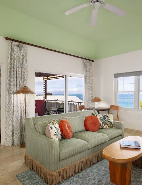 Cozy living room with a mint couch, tropical-patterned cushions, wooden coffee table, ceiling fan, and large windows opening to a sunny beach view.