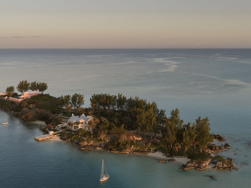 A small island with trees and a few buildings sits in clear blue water, a couple of boats anchored nearby, and a calm horizon.