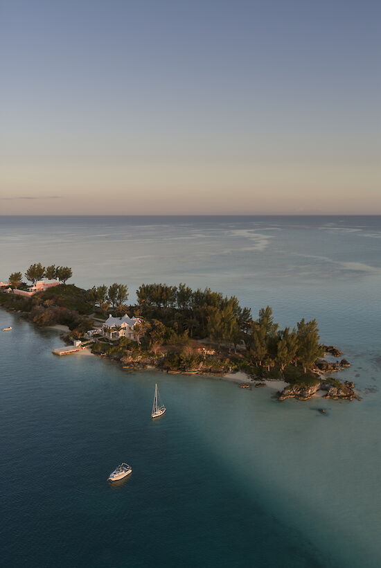 A small island with trees and a few buildings sits in clear blue water, a couple of boats anchored nearby, and a calm horizon.