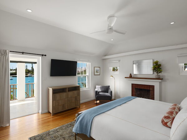 A bright hotel-like bedroom with a king bed, blue throw, fireplace, wall-mounted TV, wood floor, and balcony doors overlooking water.