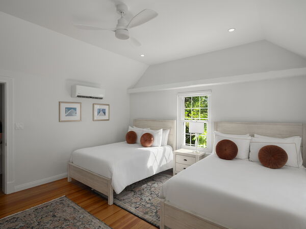 A bright, airy hotel-style bedroom with two white beds, a ceiling fan, a window, and soft gray accents; cozy decor and round brown pillows.