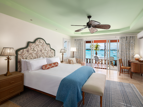A luxurious hotel room with a king bed, floral upholstered headboard, teal throw, two bedside lamps, a bench, and a balcony overlooking the ocean.