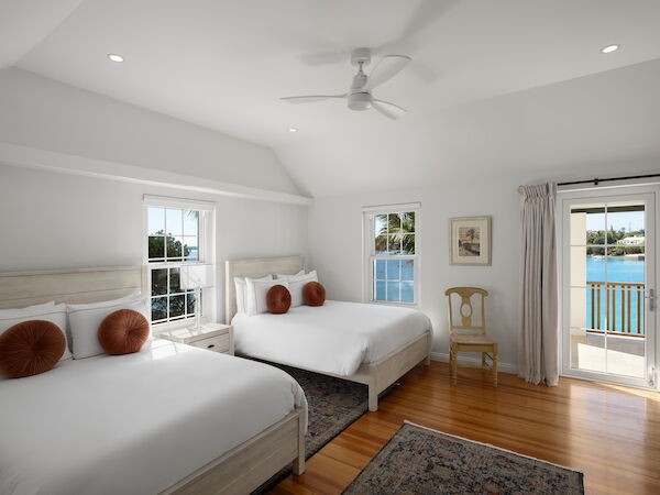 A bright coastal bedroom with two white beds, round rust pillows, a ceiling fan, light wood floors, and a balcony view to the water.