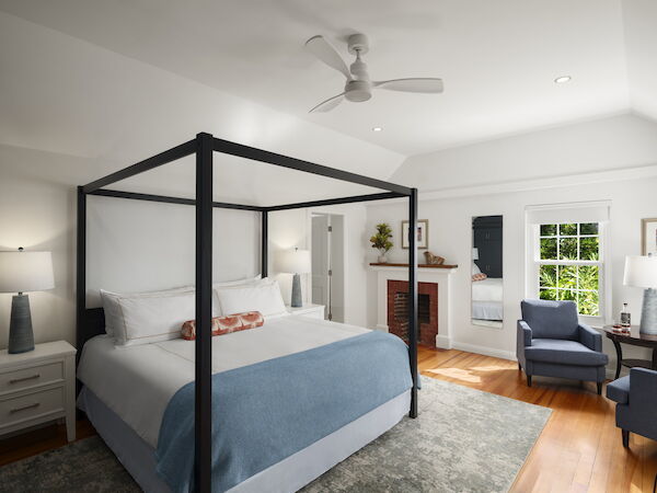 A bright hotel-style bedroom with a black four-poster bed, white walls, blue bedding, a seating area, and a cozy fireplace nook.