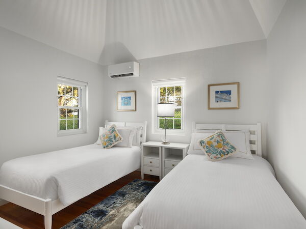 A bright, white-walled hotel room with two single beds, matching nightstands, a small rug, and a wall-mounted AC unit, sunny windows, and framed art, end.