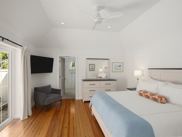 A bright, modern bedroom with white walls, a wooden floor, a king bed with light blue throw, two nightstands, a dresser, a wall TV, and a sitting area by a sliding door.
