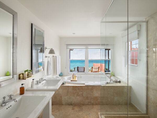 A bright hotel bathroom with dual sinks, a glass shower, and a view of an ocean-facing balcony through sliding doors.