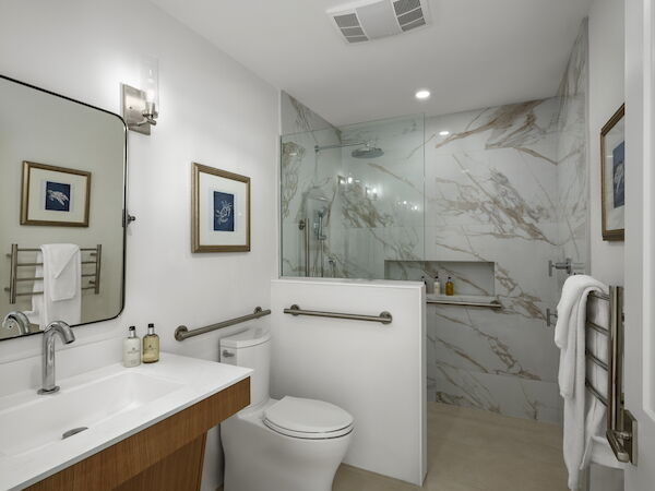 A modern bathroom with a double sink vanity, a wall mirror, framed art, a toilet, and a marble shower enclosure with a glass door, all in neutral tones.