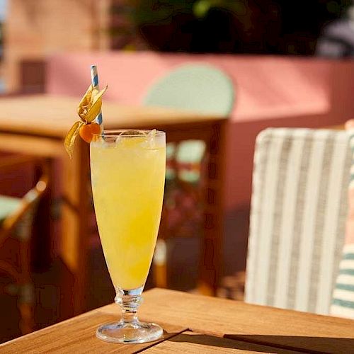 A tall glass filled with a yellow beverage and garnished with a fruit skewer is on an outdoor table with chairs and cushions in the background.