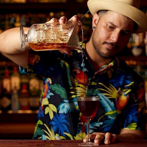 A bartender in a tropical shirt and hat pours a drink into a cocktail glass against a backdrop of a well-stocked bar.