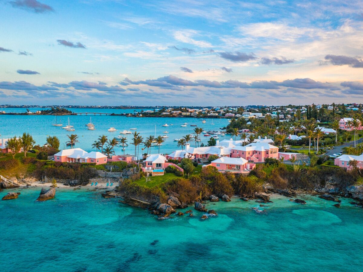 A tropical coastal town with turquoise seas, white houses, and palm trees along a rocky shoreline at sunset.