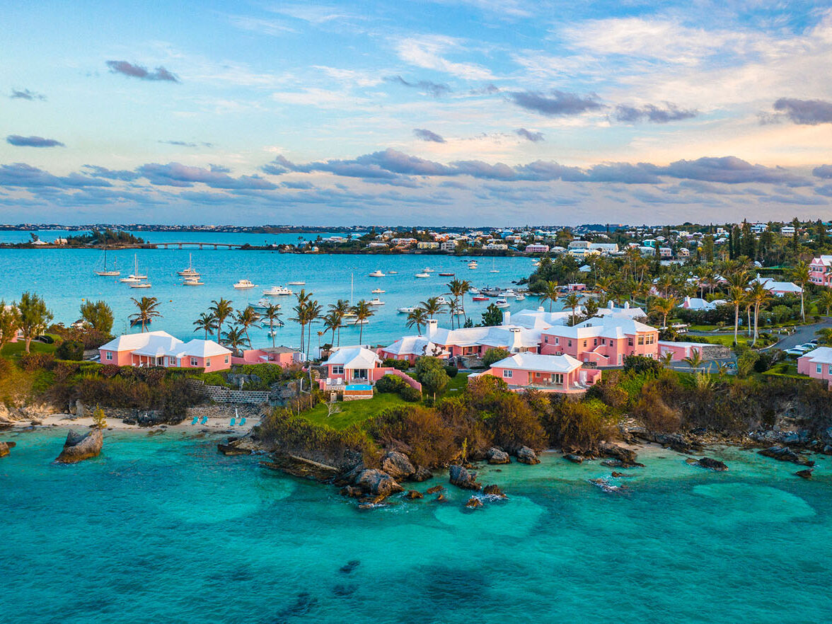 A tropical coastal scene with turquoise water, rocky shoreline, and a cluster of pink-roofed buildings along the coast under a partly cloudy sky.