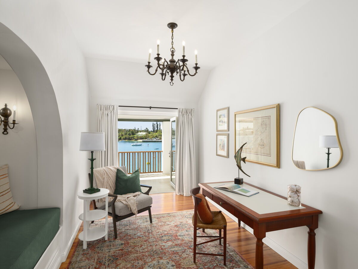 A bright hotel room with a desk, chairs, a view of the water through French doors, a chandelier, and a cozy seating nook.