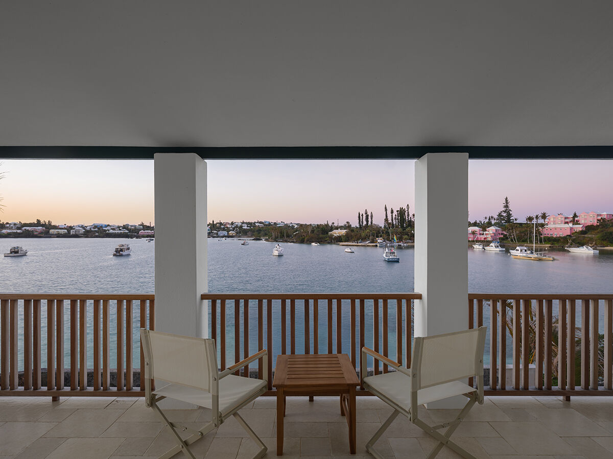 A balcony with two white chairs and a small table overlooks a calm body of water with boats; a serene waterfront scene at dusk.