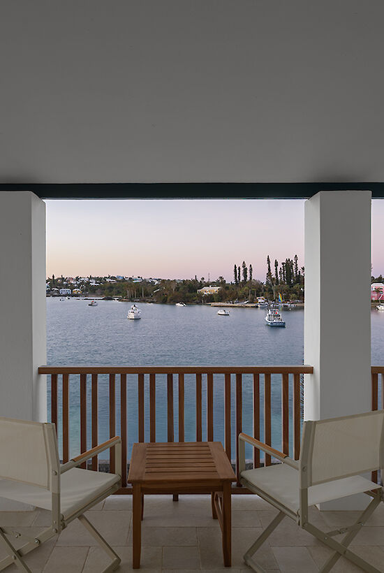 A balcony view over a calm body of water with small boats, city skyline in the distance, and two chairs with a table, under a shaded veranda.
