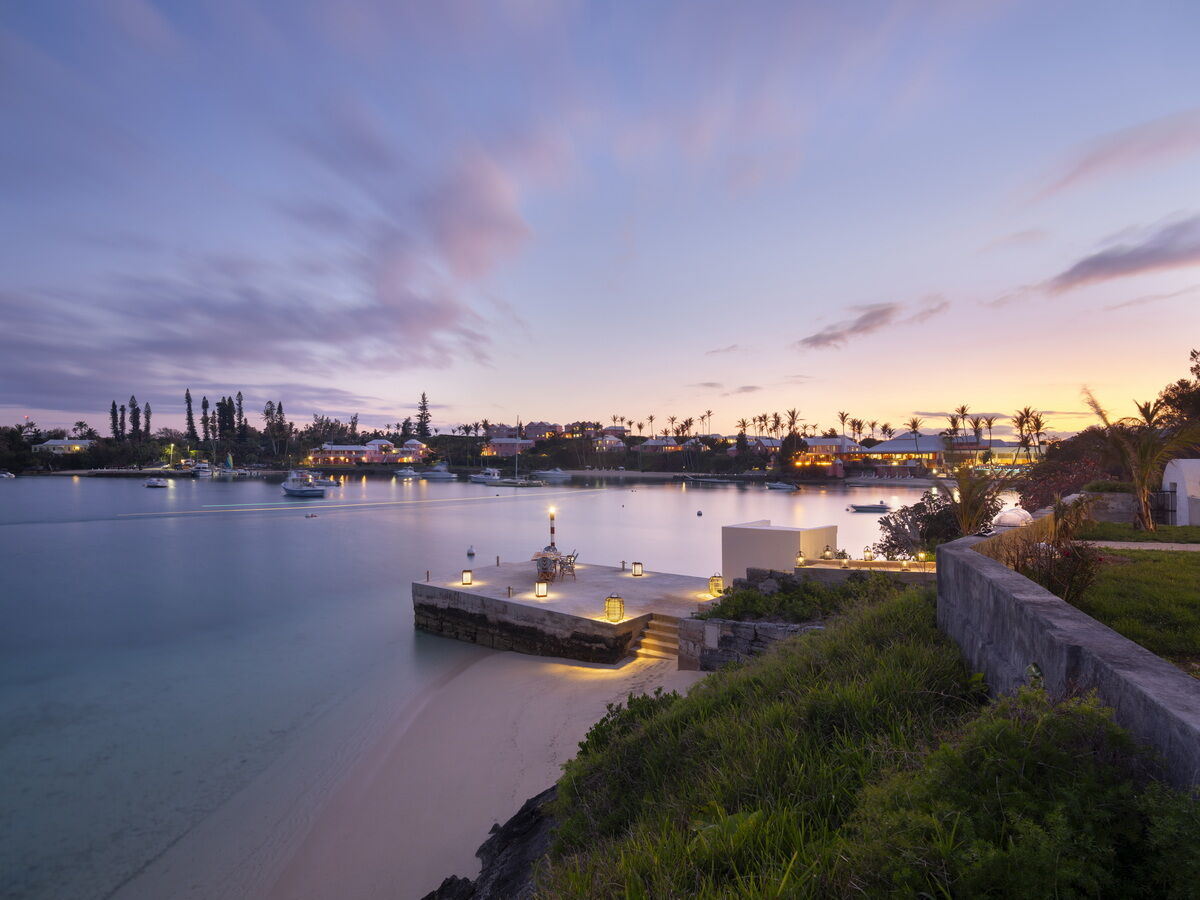 A tranquil harbor at dusk with calm water, small piers, and distant lights along a waterfront promenade, palm trees, and a pastel sky ending softly.