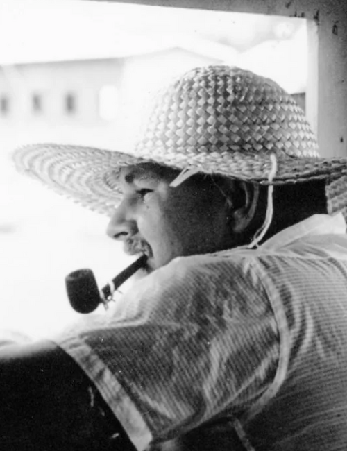 A man wearing a wide straw hat smokes a pipe, sitting by a window in black-and-white, looking off to the left.