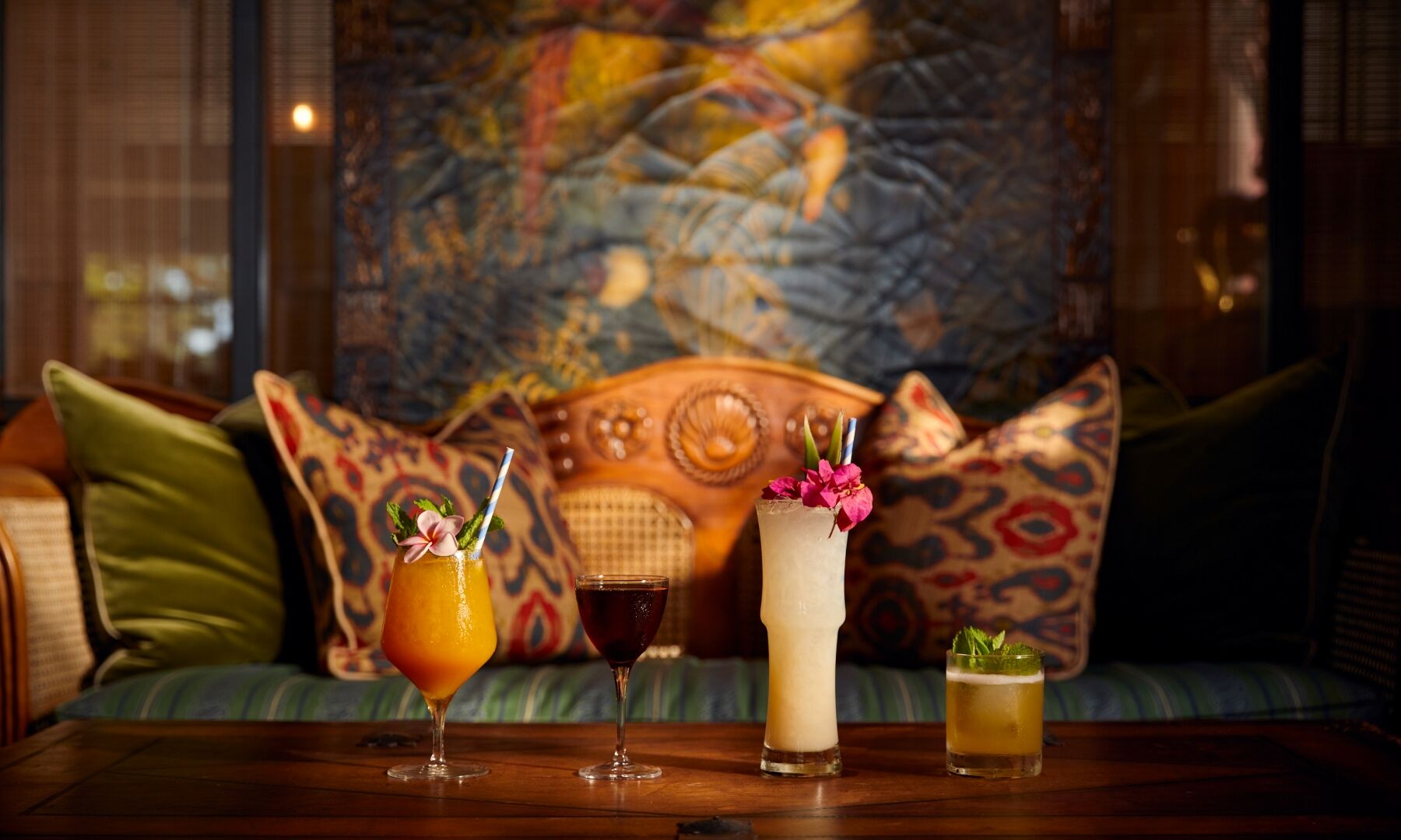 A cozy bar setup with four cocktails on a wooden table, colorful cushions and warm ambient lighting in a lounge setting.