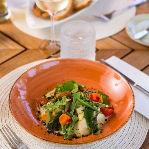 A dish of vegetables and greens on an orange plate, set on a table with a glass of white wine, cutlery, and other dining items.
