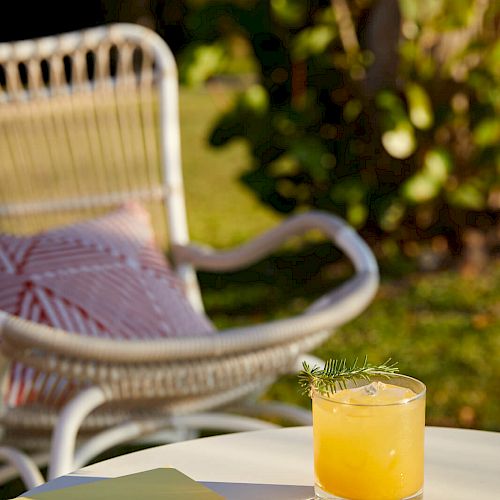 A wicker chair with a cushion, a small table with a yellow book or notebook, and a glass of orange juice garnished with herbs. Outdoor setting.