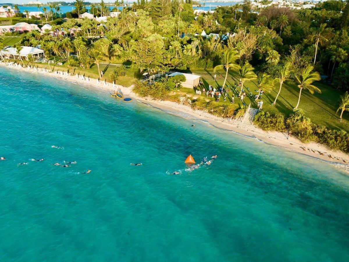 A tropical beach with turquoise water, white sand, and palm-lined shore; a small boat or buoy floats near the shore, with greenery and buildings in the background.