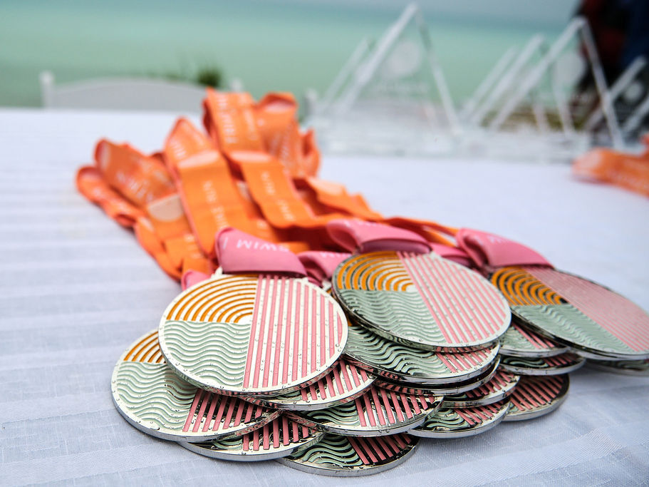 A pile of striped round medals with orange ribbons on a white table, likely award medals for a competition, on a beach setup.