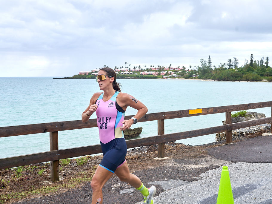 A female runner wearing pink and blue athletic gear jogs along a coastal road by a wooden fence near the water, with a cone nearby.