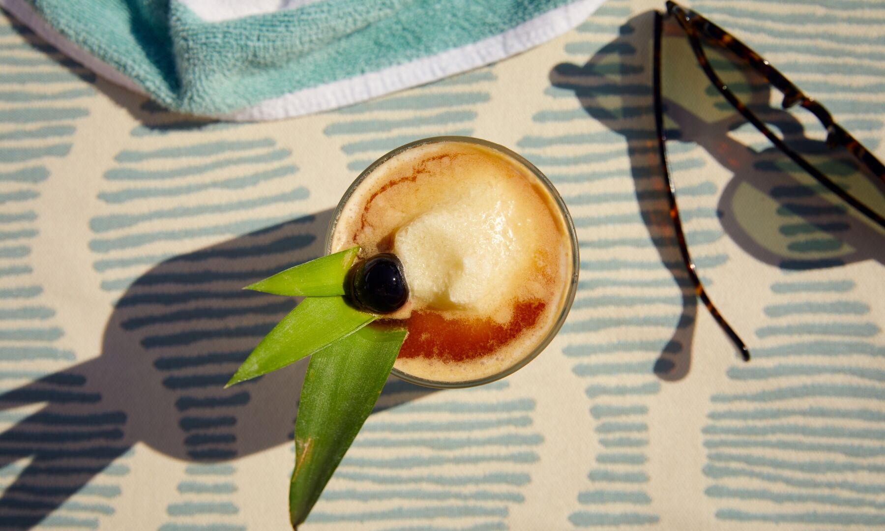 A tropical drink with a cherry and lime garnish on a patterned table, beside sunglasses and a towel. All set for a sunny afternoon.