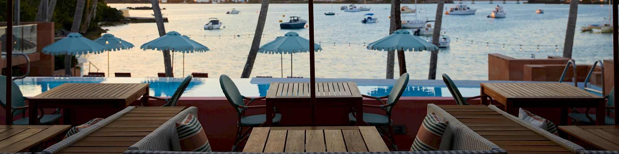 The image shows a scenic coastal area with tables and chairs under green umbrellas by the water, with boats and palm trees visible in the background.