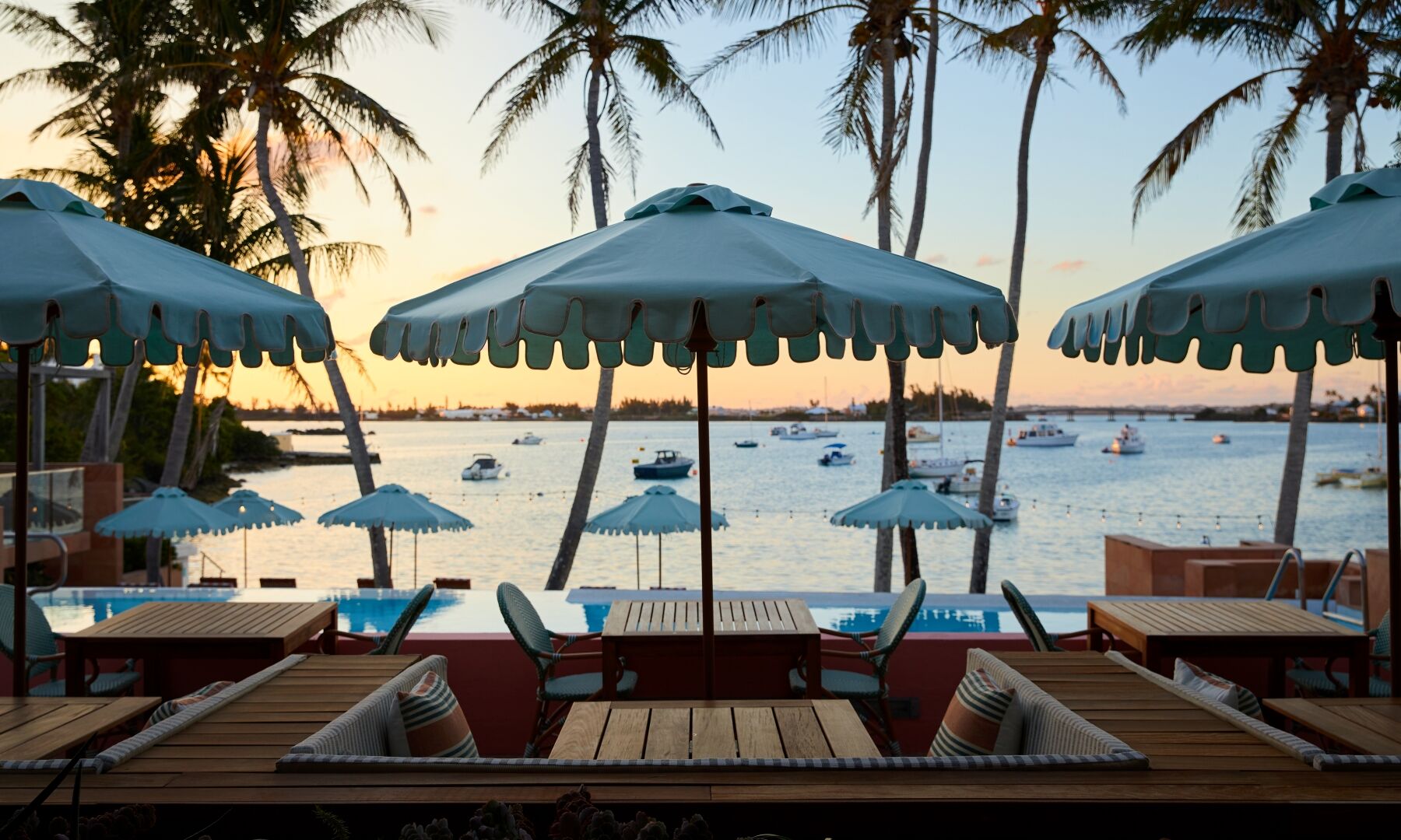 Beachside lounge with wooden tables, blue umbrellas, and palm trees at sunset; calm water and boats in the distance.