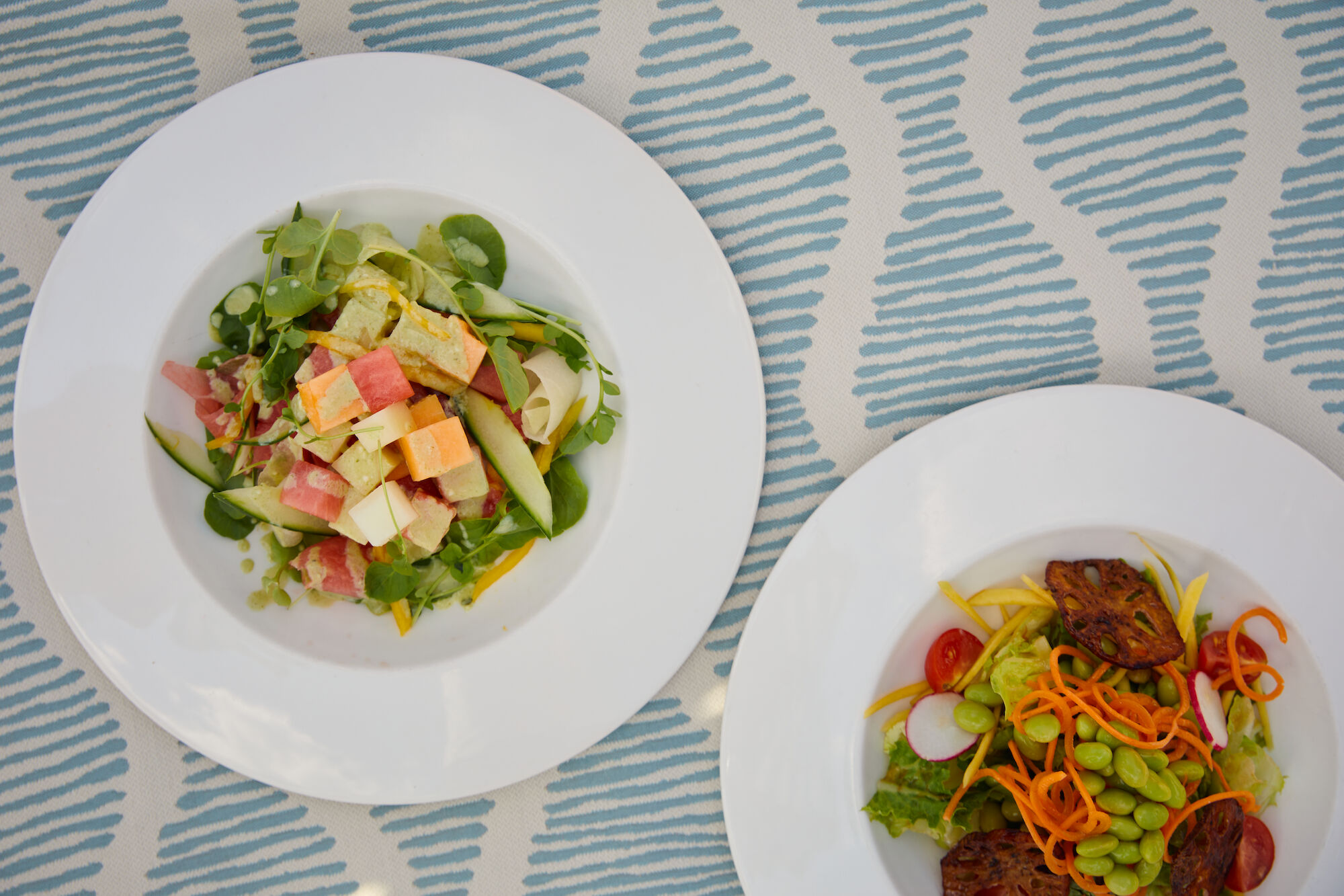Two white plates on a patterned blue table: left plate has a mixed salad with greens and cubed ingredients; right plate features noodles with vegetables and meat.