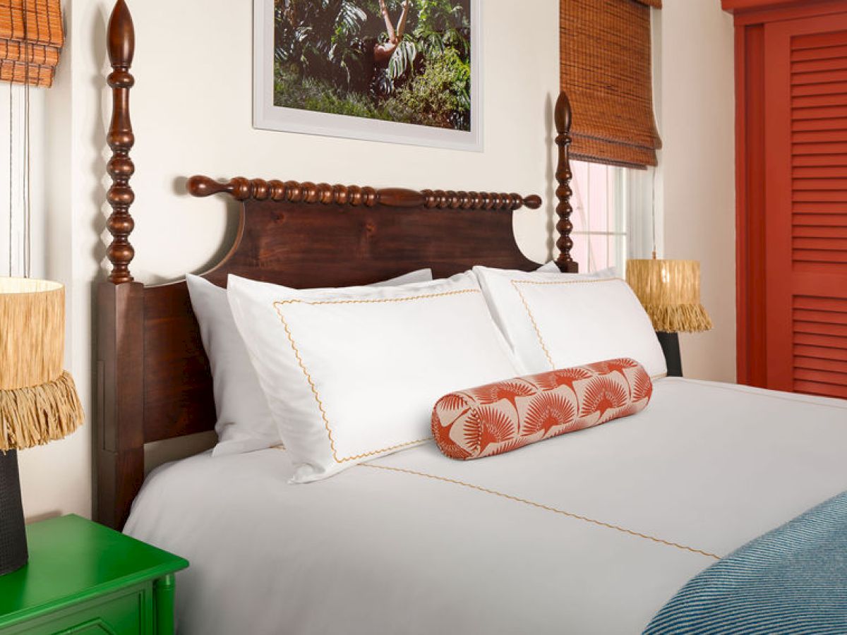 A cozy bedroom with a wooden bed, white linens, patterned pillows, green nightstand, wicker lampshades, wall art, and bamboo window blinds.