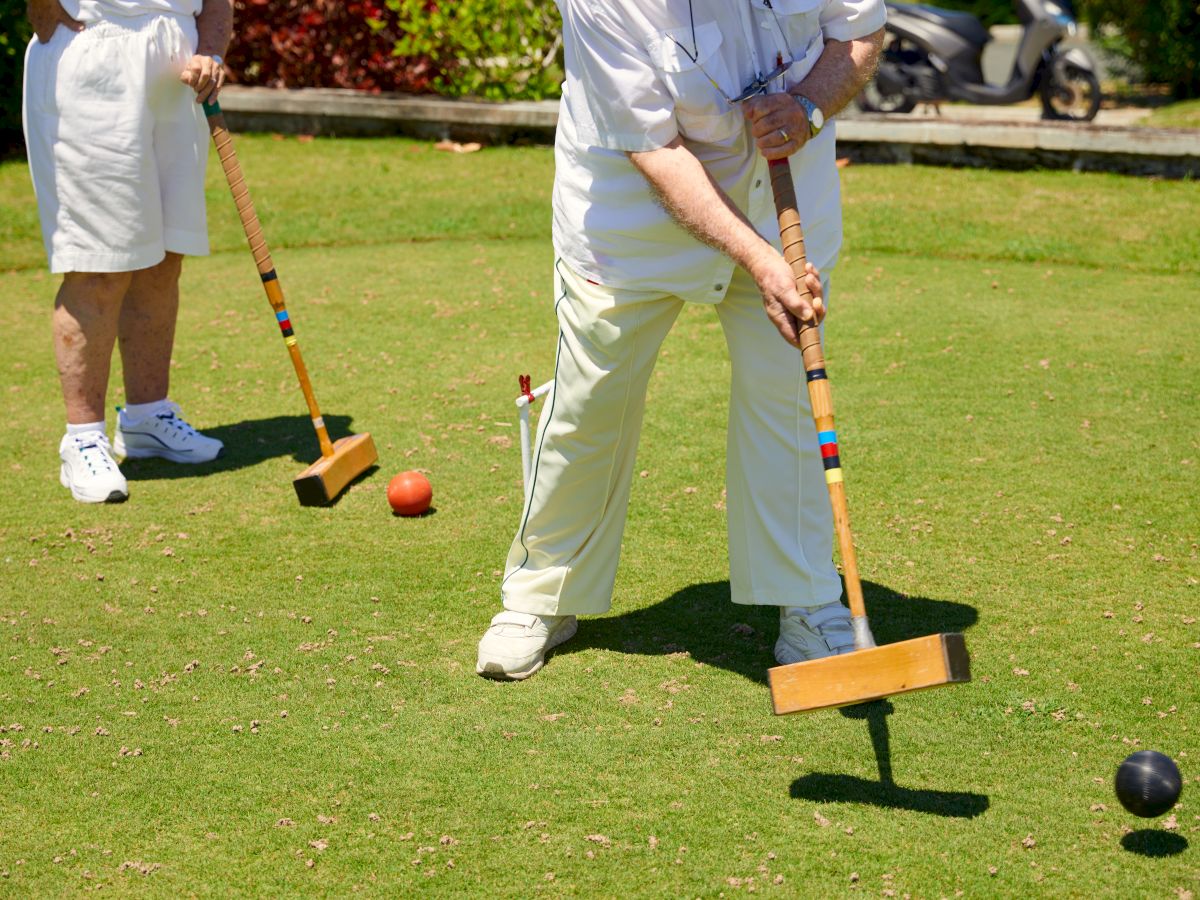 Two people are playing croquet on a grassy lawn. Both are dressed in white, holding mallets, aiming to hit colored balls through wickets.
