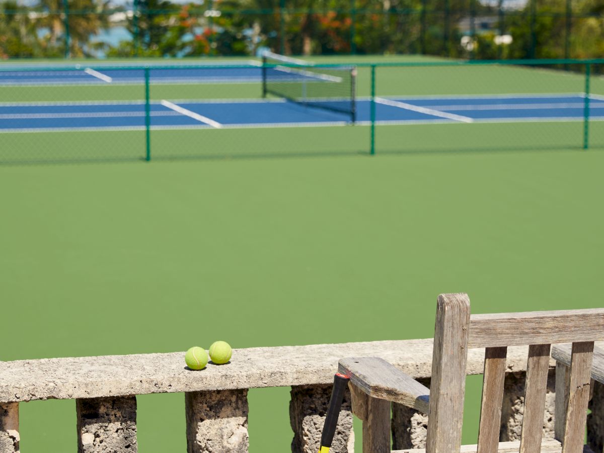 A sunny tennis court with a bench, tennis balls, and a racket placed next to it. Trees and a blue sky are in the background.