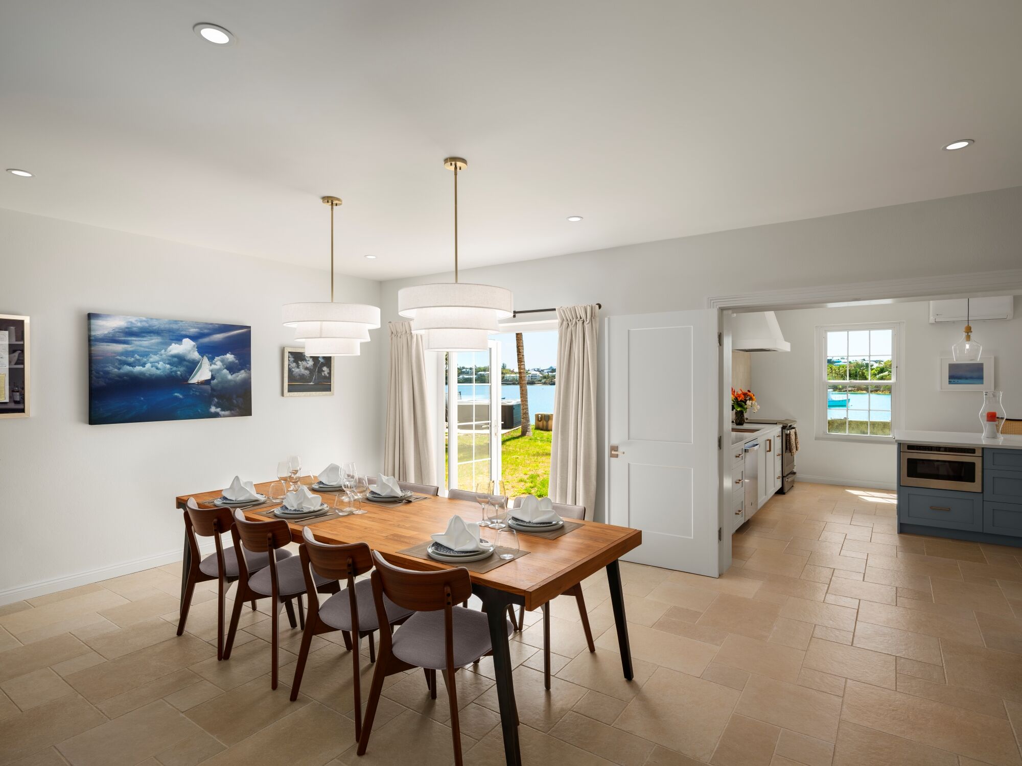A bright, modern dining room with a wooden table for six, tan tiles, white walls, and a view into a sunny kitchen; minimalist decor.