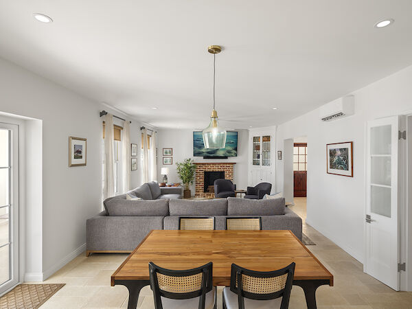 A bright, modern living room with a gray sectional, wood dining table in the foreground, a fireplace, and a mounted TV across from the seating area.