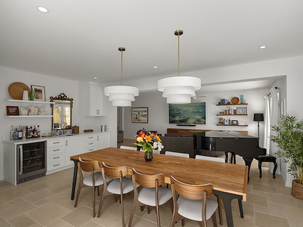 A bright, open dining room and living area with a wooden table for eight, white cabinetry, floating shelves, a piano, and a large flat-screen TV.