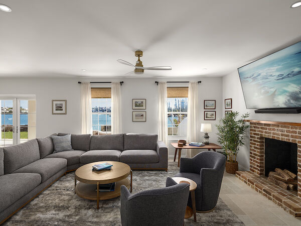 A cozy living room with a large gray sectional, two chairs, round coffee table, brick fireplace, a big wall-mounted TV, and light streaming through multiple windows.
